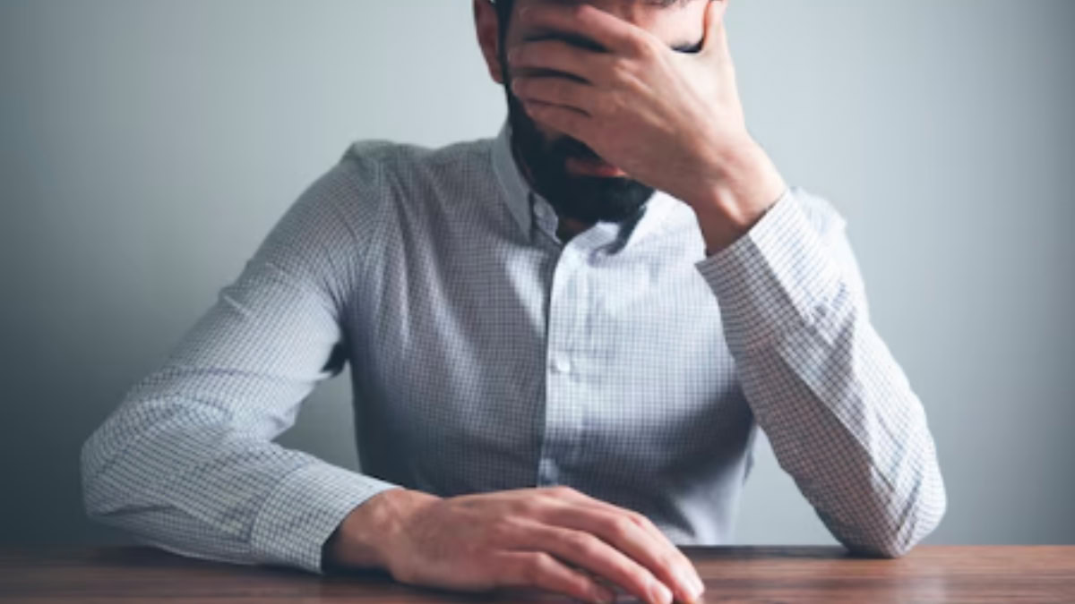 Homem sentado à mesa com expressão de angústia e a mão no rosto, simbolizando os efeitos emocionais da ansiedade e o impacto no cotidiano.