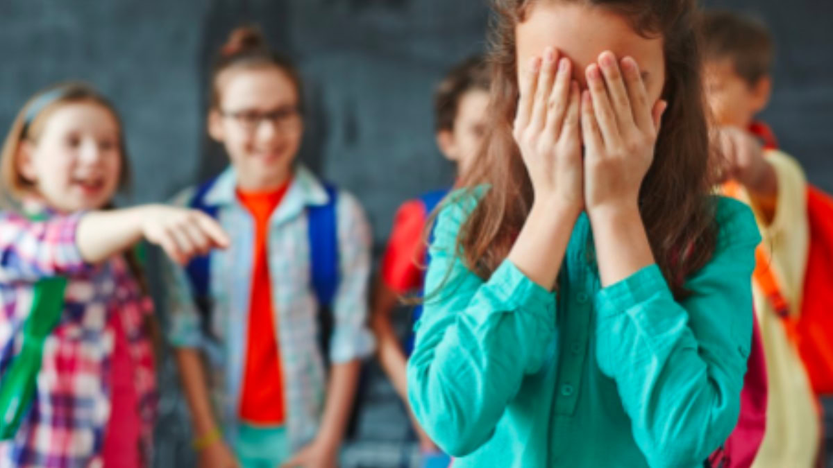 Menina em idade escolar cobrindo o rosto com as mãos enquanto outras crianças riem e apontam para ela ao fundo, representando sofrimento emocional na infância e bullying.