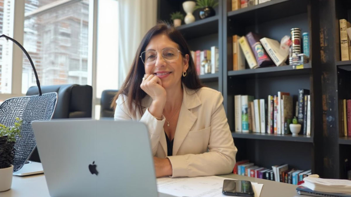 Psicóloga Genori Oliveira sorrindo em atendimento online, sentada à mesa com notebook aberto em um ambiente acolhedor com estante de livros ao fundo.